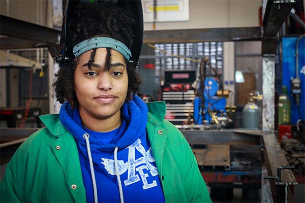 A high school student wearing a welding helmet with the mask uplifted stands in a workshop at the Hub.