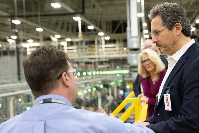 Three people in safety glasses and a fourth person partially visible in the background talk as they look over a production floor.