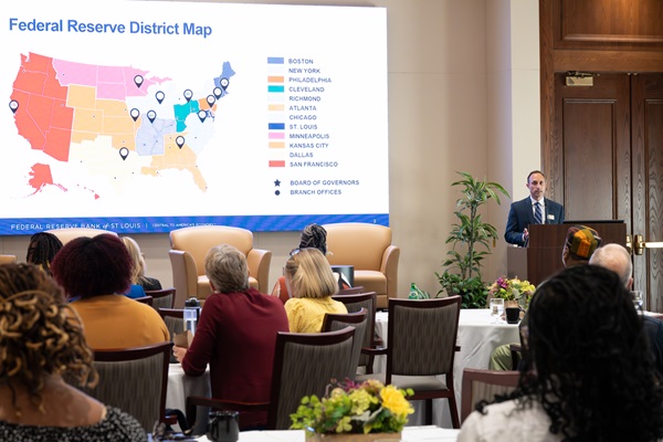 A man in business attire speaks from a podium to a group with a large screen behind him showing a Federal Reserve district map.