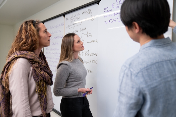 Two women on the left of the frame and one man look at a whiteboard with equations on it.