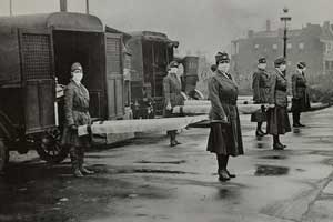 Mask-wearing women hold stretchers at the backs of ambulances.