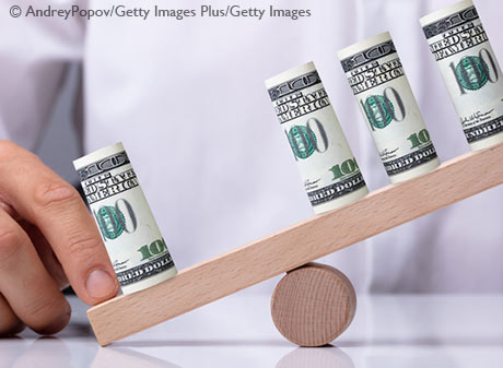 Rolls of U.S. currency are on top of a small wooden seesaw, which is kept off balance by a human hand.