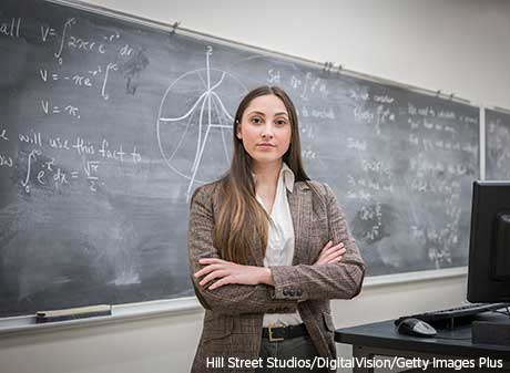 A person, with arms crossed, stands before a blackboard in a classroom.
