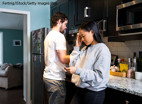 A young woman views a financial document in her apartment while a friend is cooking a meal.