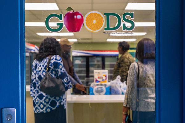 Four people are seen through a glass door with 'GOODS' lettering on it. The people are standing around  a counter with groceries on it in front of cooler cases.