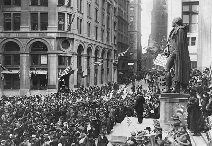 A black and white photo shows downtown streets thronged with people, U.S. flags and a statue with “Liberty Forever” hung from it.