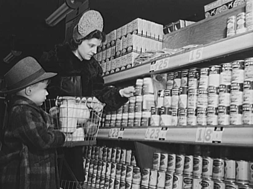 A black and white photo shows a woman and little boy in 1940s attire shopping for canned goods.