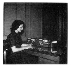 In a black and white photo, a woman in business attire operates a desktop machine.