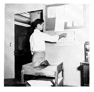 In a black and white photo, a man in business attire moves a card on a wall-mounted holder.