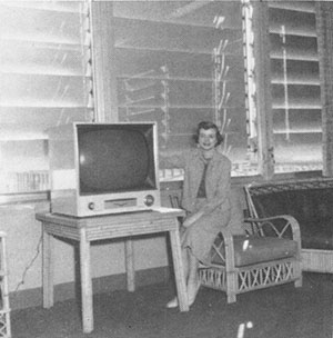 In a black and white photo, a woman in a business suit perches on chair arm next to a boxlike TV on a table.