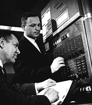 In a black and white photo, two men in business suits work on a computer.