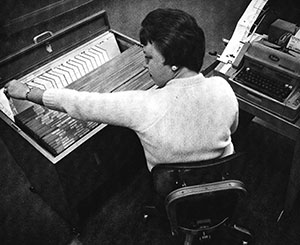 In a black and white photo, a woman in business attire sits and operates one of two desktop machines.