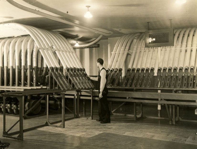 In a black and white photo, a man in a tie and vest holds one tube in a roomful of tubes.