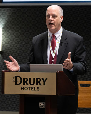 Man in suit and tie speaks at a hotel lectern.'