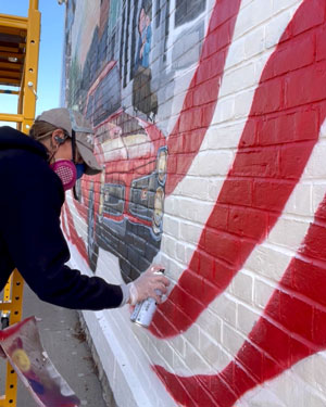 Woman in baseball hat and paint respirator spray paints a flag stripe on a brick wall mural.'