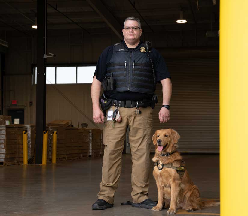 Law enforcement officer stands in warehouse doorway next to golden retriever.