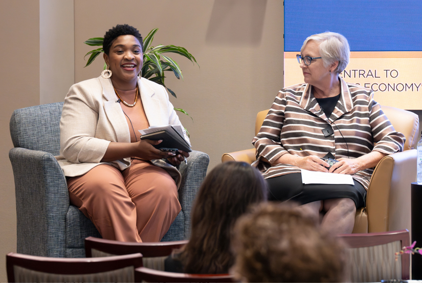 A woman in business attire speaks from an armchair at the front of a conference room while another woman seated next to her looks on.