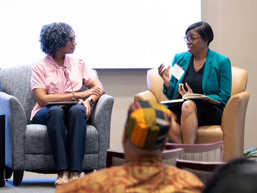 Two women in business attire sit side by side in armchairs; one speaks while the other looks on.