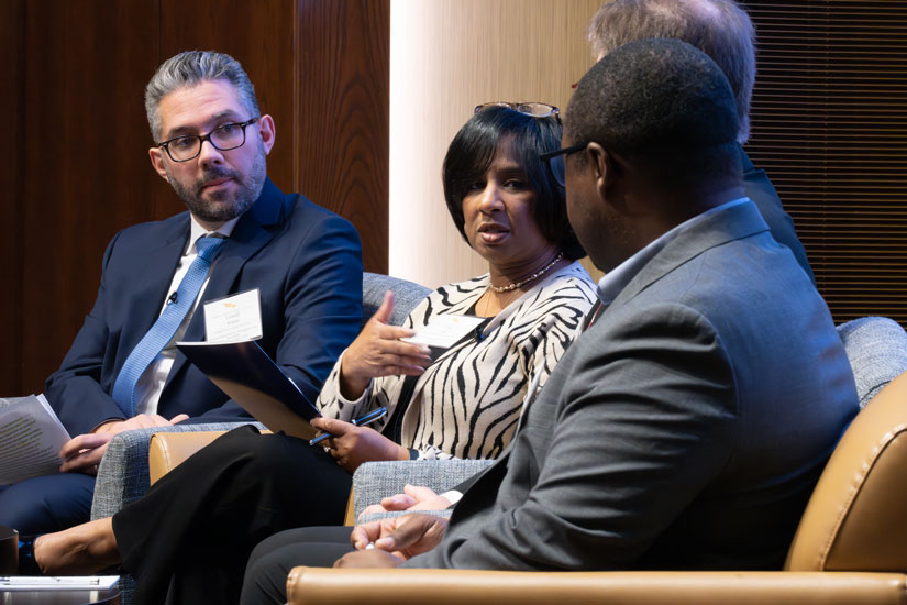 A moderator and three panelists in business attire sit in a row of armchairs at the front of an auditorium.