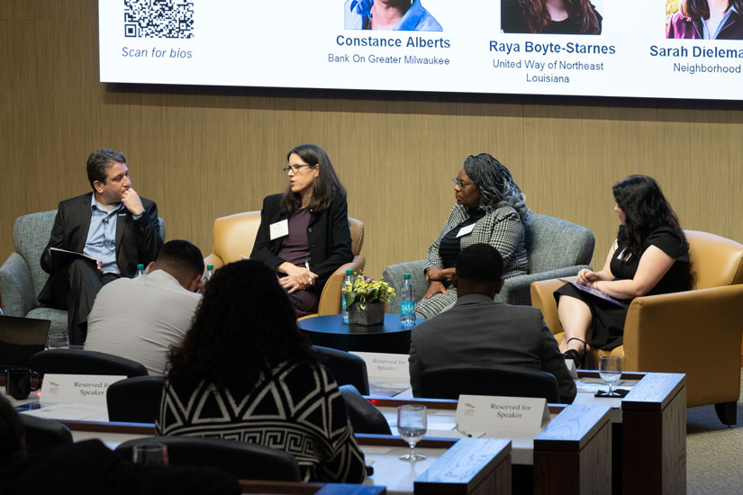 A moderator and three panelists in business attire sit in a row of armchairs at the front of an auditorium facing their audience.
