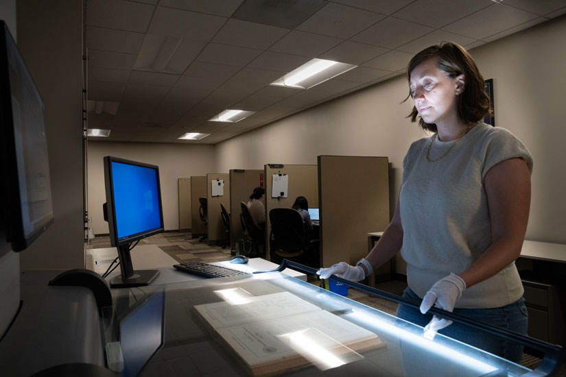 A woman wearing white gloves with her face illuminated uses a machine to digitize material from a book while people work in cubicles in the background.