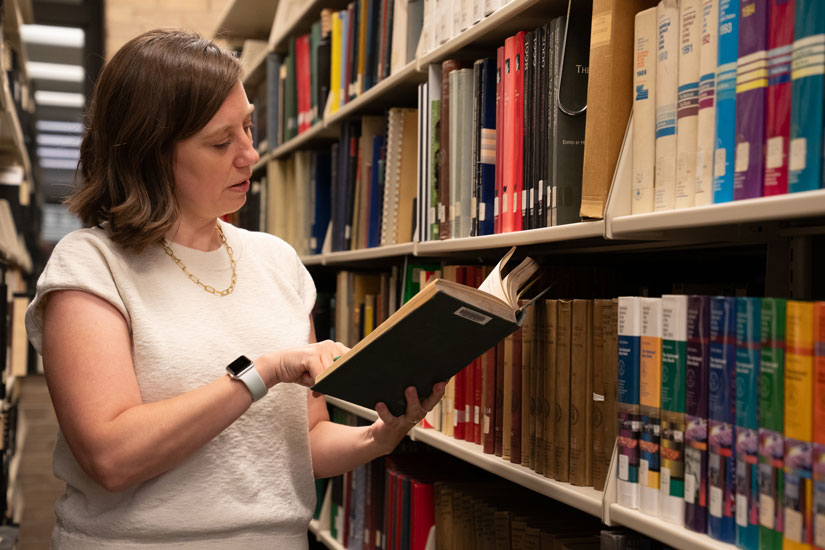 A person holds an open book while standing next to shelves of books.