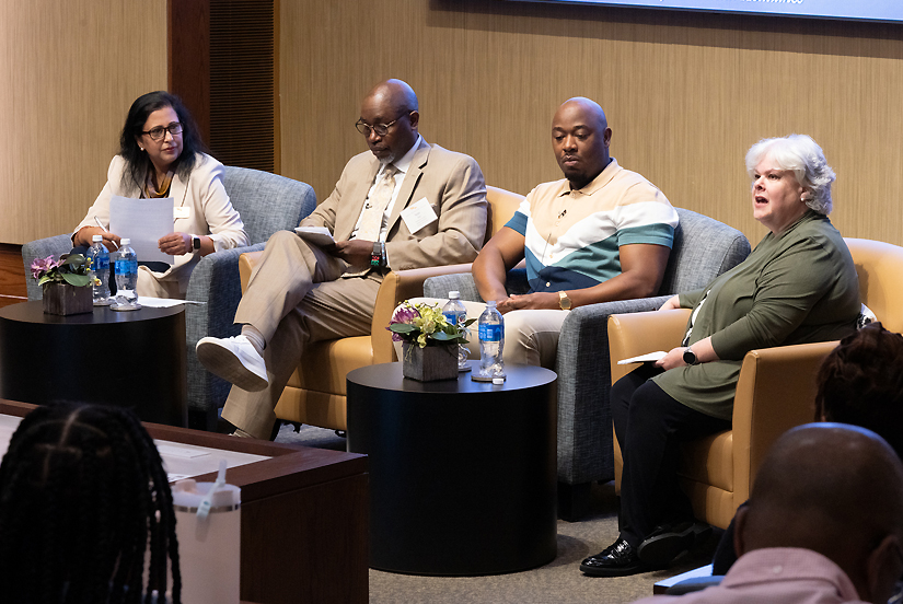 A moderator and three panelists in business attire sit in a row of armchairs at the front of an auditorium.
