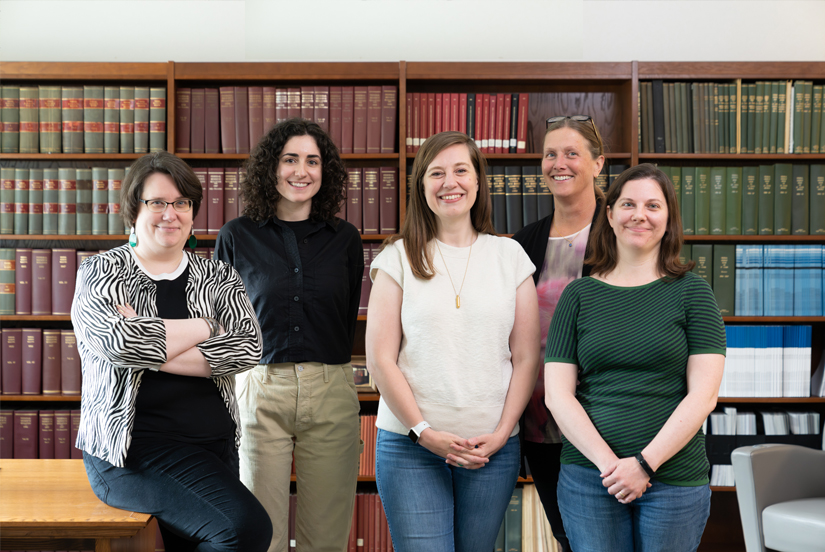 Five women in business casual attire pose in front of shelves of book collections.