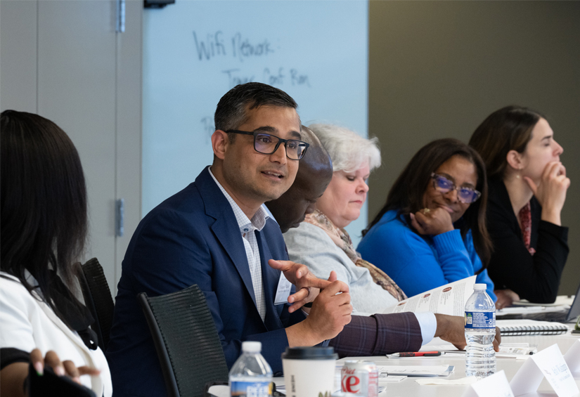 A person seated at a conference table with five others speaks while the others listen.