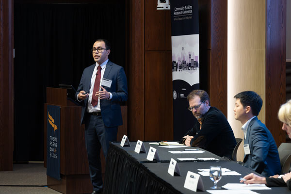 A man in business attire speaks at a lectern next to three people in business attire at a table.