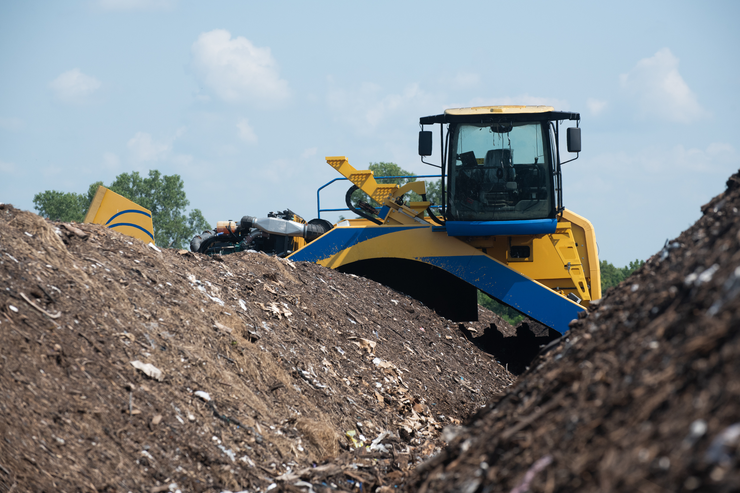A yellow and blue machine with driver’s seat visible through the windshield sits in heaps of earthen material.