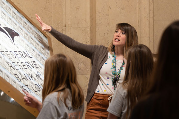 A woman in front of a group of children gestures at a glass cube with currency inside and “Federal Reserve Bank of St. Louis” printed on it.