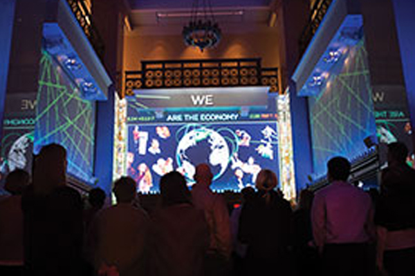 Wall screens show a video with “We Are the Economy” text on three walls of a darkened room filled with standing people.