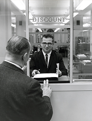 Man at teller window with “Discount” on the glass talks with a client.