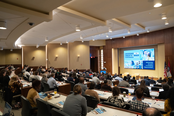A full auditorium has a slide displayed on a screen in front with photos of families and the text ‘Crossing the Credit Barrier conference.’