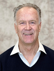 Man in a collared shirt and sweater smiles in front of a gray background.