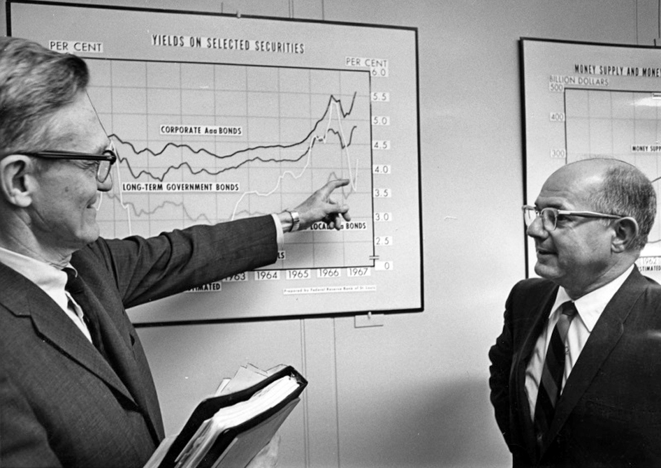 A man in business attire and glasses gestures at a line graph on a wall while talking with another man in business attire and glasses.