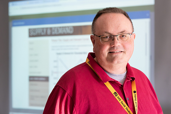 A man wearing a red polo shirt and a lanyard stands in front of a screen.