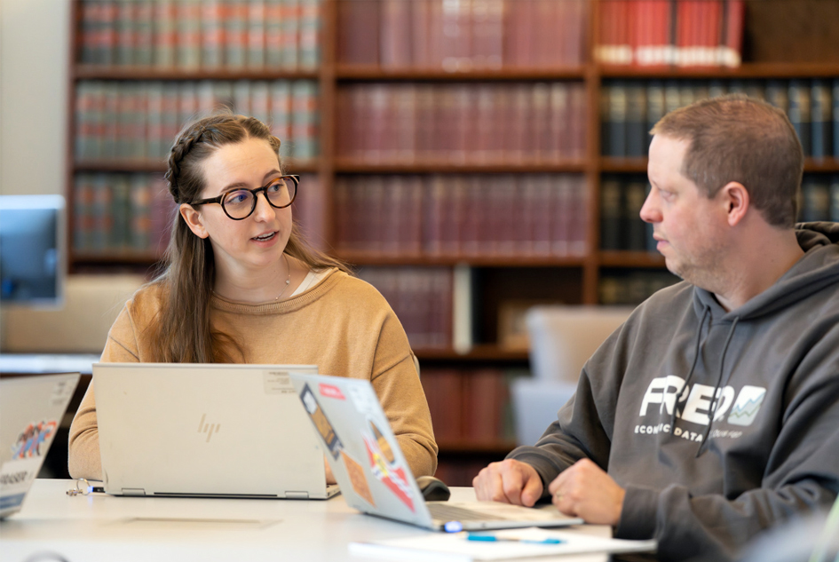 Two people sit with laptops at a table in front of shelves of books.