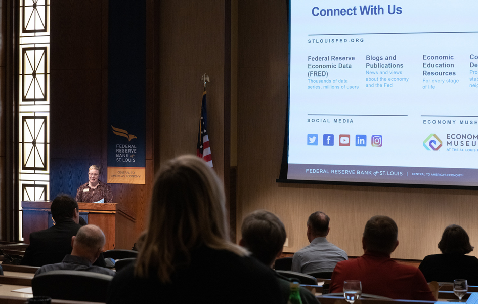 An audience in an auditorium listens to a person behind a lectern with a screen showing a slide with ‘Connect With Us’ text.