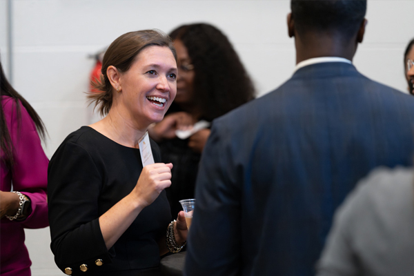  A woman in business attire smiles while talking with a man in business attire at a gathering.