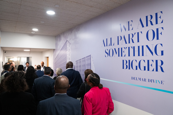  A group of more than a dozen people in business attire listen to two speakers while standing in a hallway that has images of buildings and the text “We are all part of something bigger. Delmar Divine” painted on a wall.