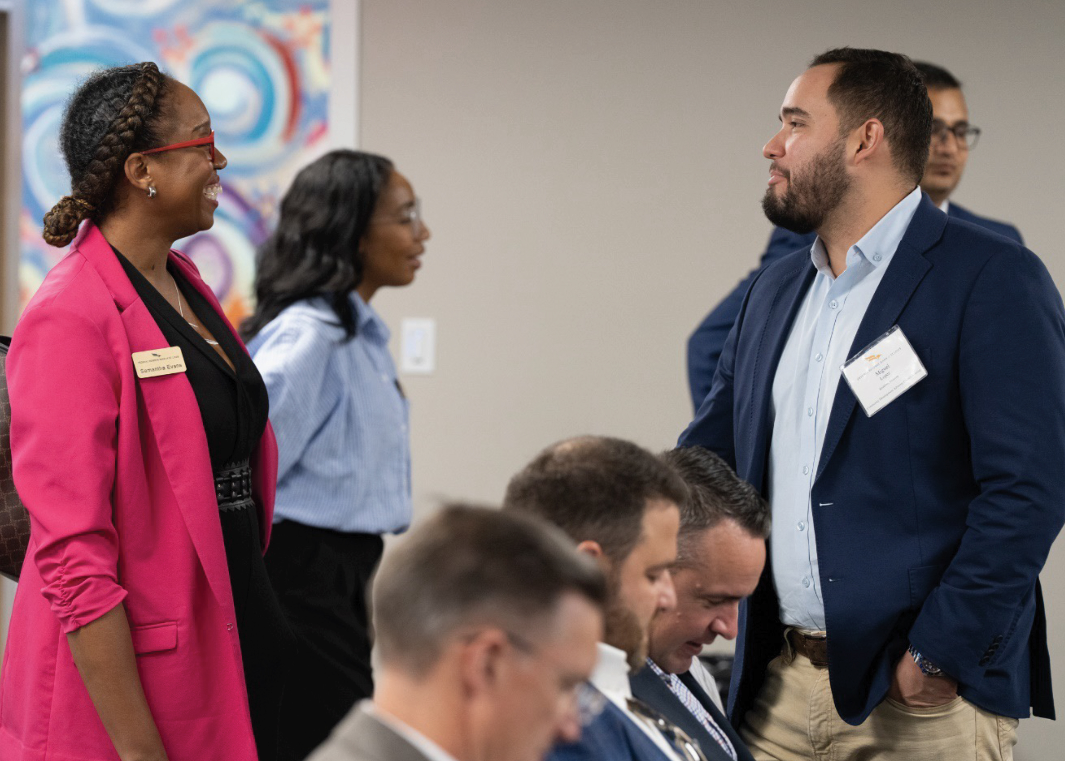 A woman in business attire smiles while talking with a man in business attire at a gathering.