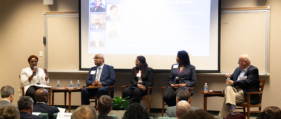 An audience watches as a woman sitting on a stage with four other people speaks into a microphone. A screen shows photos behind the stage.
