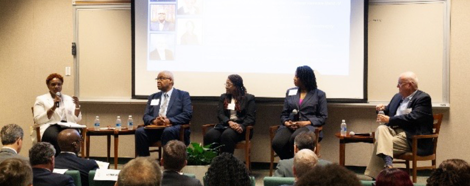 An audience watches as a woman sitting on a stage with four other people speaks into a microphone. A screen shows photos behind the stage.