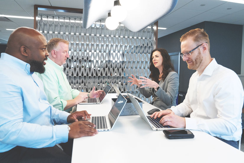 Three men and one woman in business attire sit at a four-person table with their laptops engaged in positive discussion.