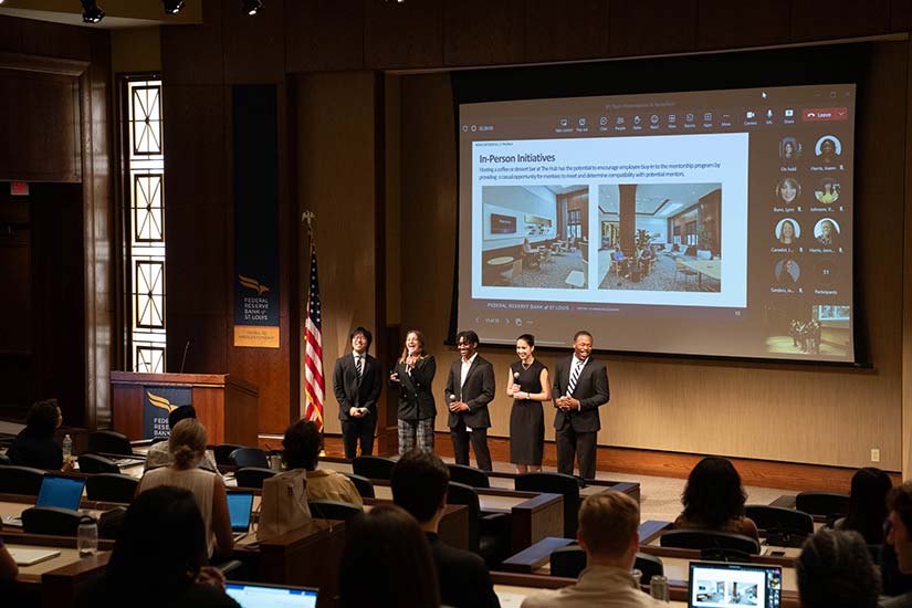 Five students presenting to an auditorium filled with people in front of a large screen.