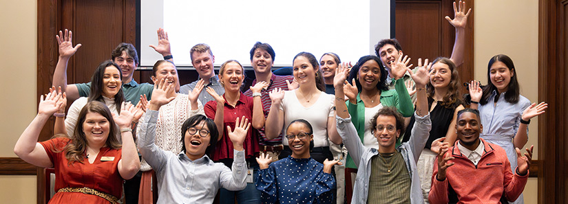 Group of interns and employees in brightly colored shirts smile and raise their hands in the air, with a projection screen in the background.