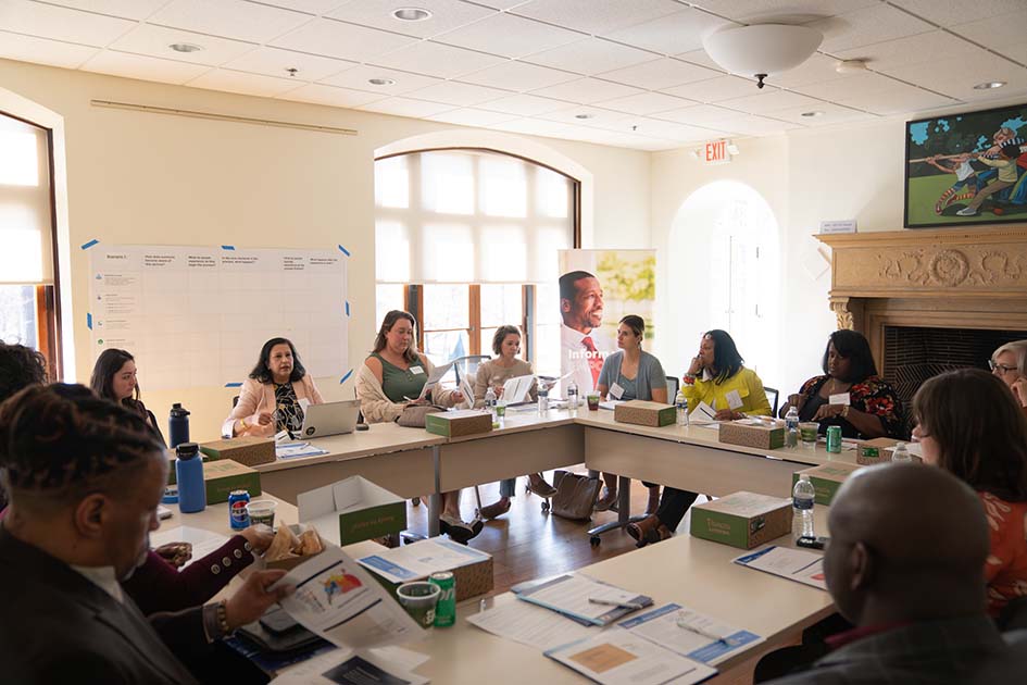 A group of people with nametags and presentation materials sit and converse around a square made by tables in a bright, sunlit room.