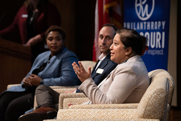 A woman speaks while two co-presenters turn toward her to listen.